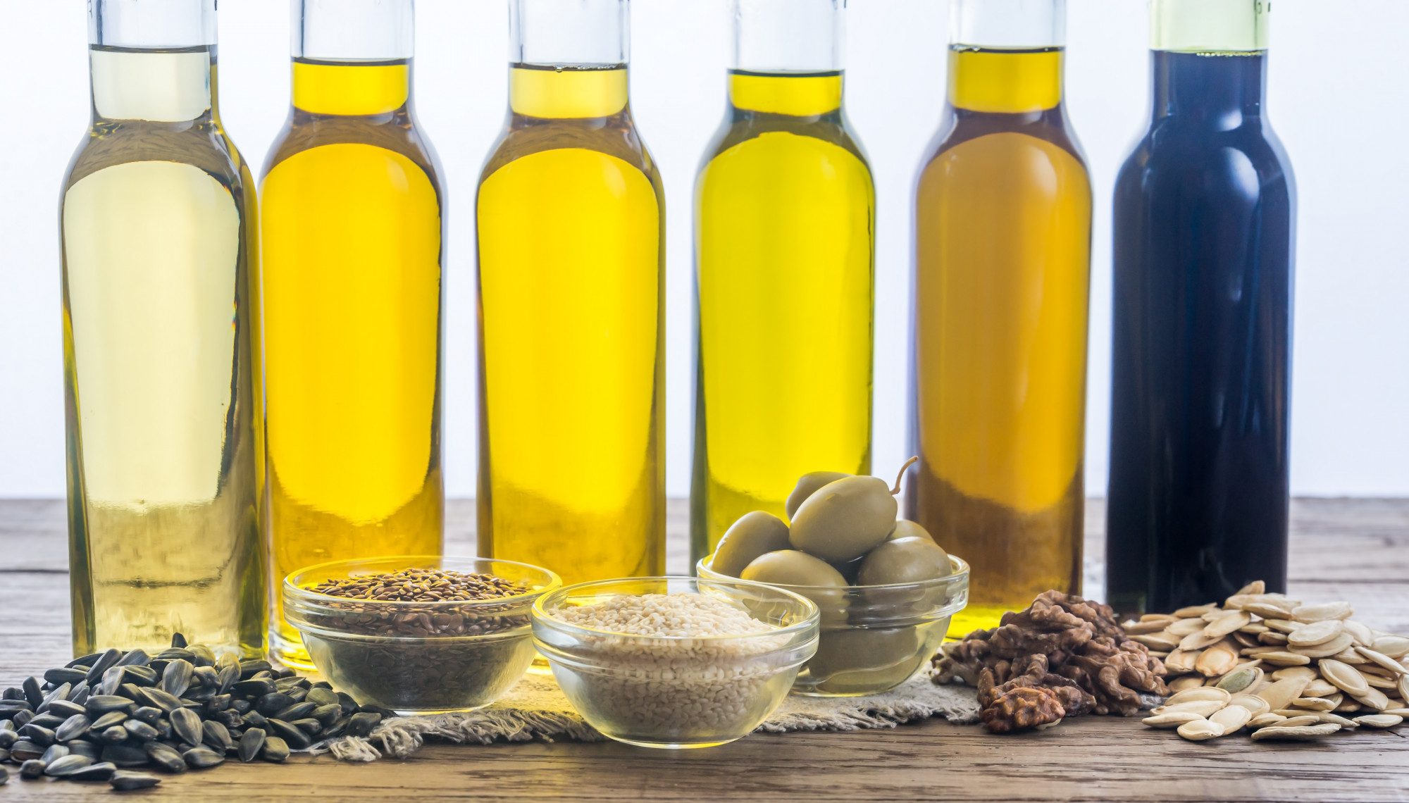 Assorted cooking oils including CanolaMAX, with seeds, grains, and olives on a wooden table against a white background.