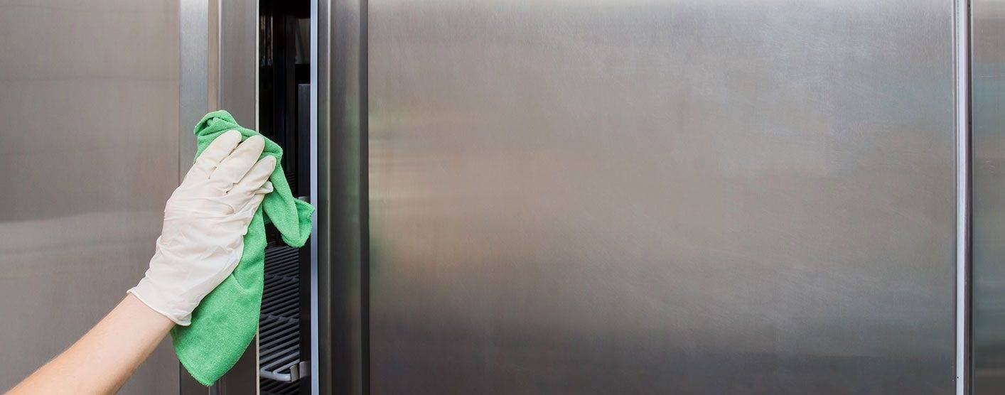 A person wearing gloves uses a green cloth to wipe down stainless steel surfaces, highlighting the importance of deep cleaning commercial kitchens.